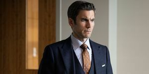 Ο Wes Bentley με ένα σκούρο γκρι κοστούμι τριών κομματιών με μια καφέ στάμπα γραβάτα και λευκό και μπλε ριγέ πουκάμισο στέκεται σε ένα γραφείο με τα χέρια στις τσέπες