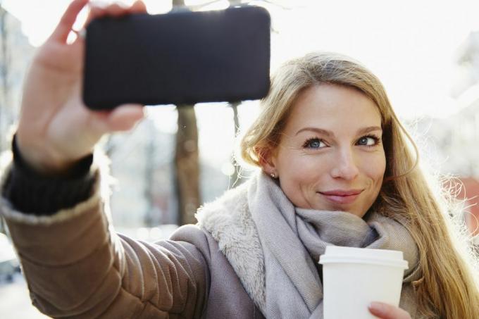 όμορφη γυναίκα που περπατά στους δρόμους της πόλης το χειμώνα βγάζοντας μια selfie και κρατά έναν καφέ για να πάει, με οπίσθιο φωτισμό