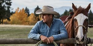 Ο kevin Kostner στο yellowstone δίπλα σε ένα άλογο ακουμπισμένο σε έναν φράχτη με σχοινί στα χέρια φορώντας ένα ξεθωριασμένο μπλε τζιν πουκάμισο και μπεζ καουμπόικο καπέλο