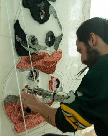 Γενειοφόρος 30χρονος Ισπανολατίνος φορώντας φανέλα ράγκμπι, στο εργαστήριό του δουλεύοντας σε χειροποίητα χαλιά φτιαγμένα με όπλο και ακρυλικές ίνες, με σχέδιο κουβέρτας σκύλου